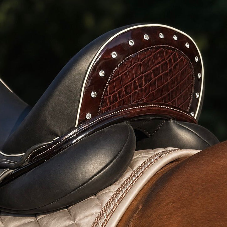 Close-up of a horse saddle resting on a horse's back, showing black leather, dark brown textured leather, silver studs, and a beige quilted saddle pad against a blurred green outdoor background.