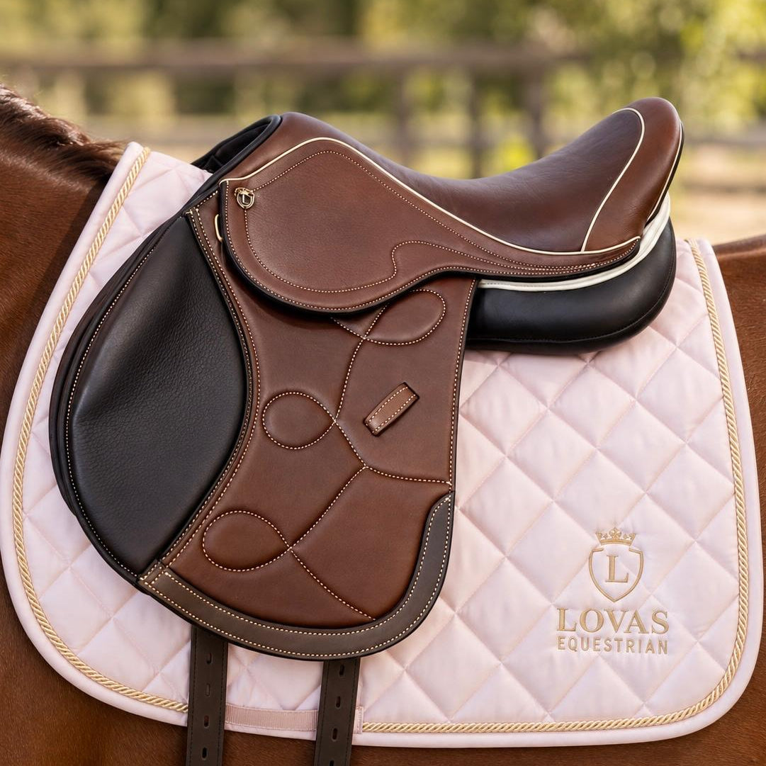 A brown leather equestrian saddle with black panels and white piping, resting on a light pink quilted Lovas Equestrian saddle pad over a brown horse, with a blurred outdoor background.