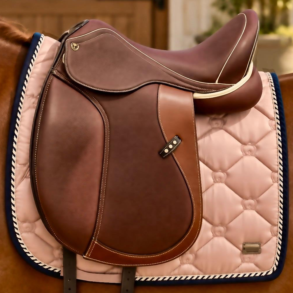 A brown leather horse saddle resting on a pink quilted saddle pad with a navy and white braided edge, shown on a horse in an indoor stable setting.