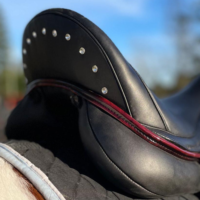A close-up shot of a black leather equestrian saddle with silver studs, resting on a dark gray quilted saddle pad atop a horse's neck blanket, with a blurred outdoor background of blue sky and trees.