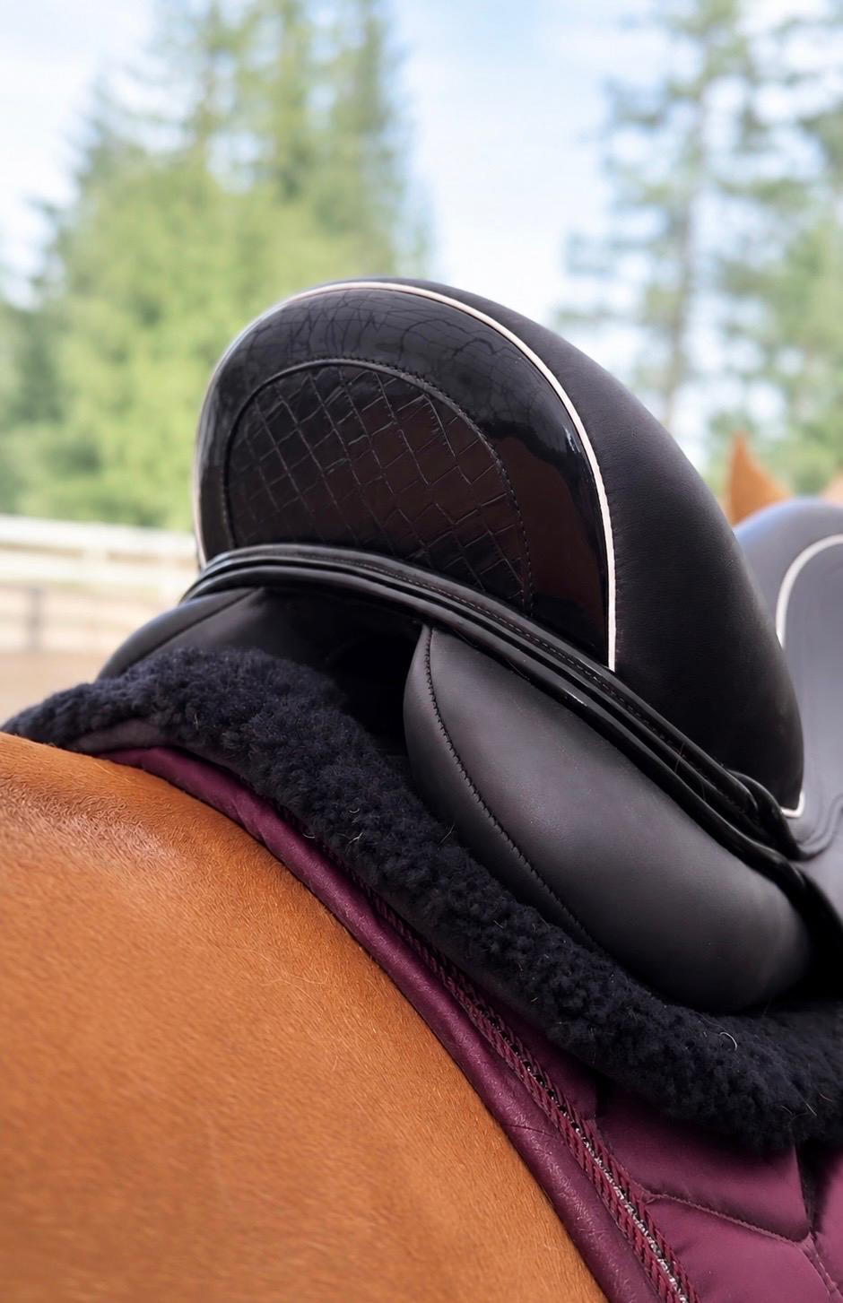 Close-up of a black leather horse saddle with a dark fleece or shearling lining, positioned on a horse's brown mane over a burgundy saddle pad, with a blurred outdoor background of trees and sky.