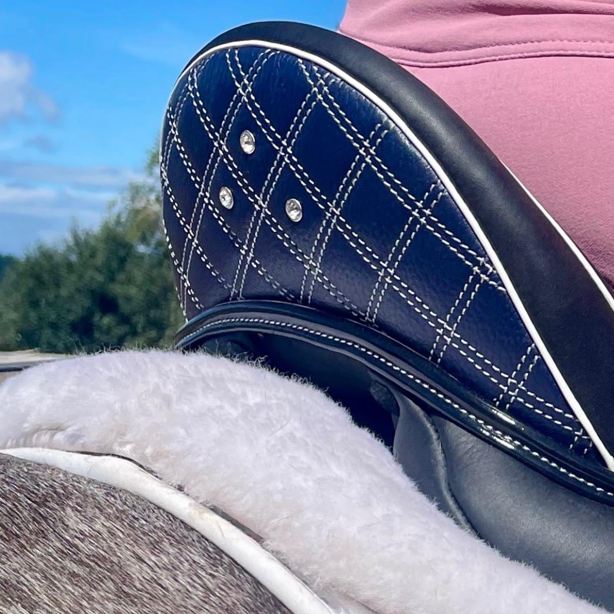 Close-up of a horse saddle pad with a navy blue quilted leather top, white stitching, rhinestone accents, a white fleece layer, and a brown horse's hair underneath, with a pink saddle blank visible in the background.