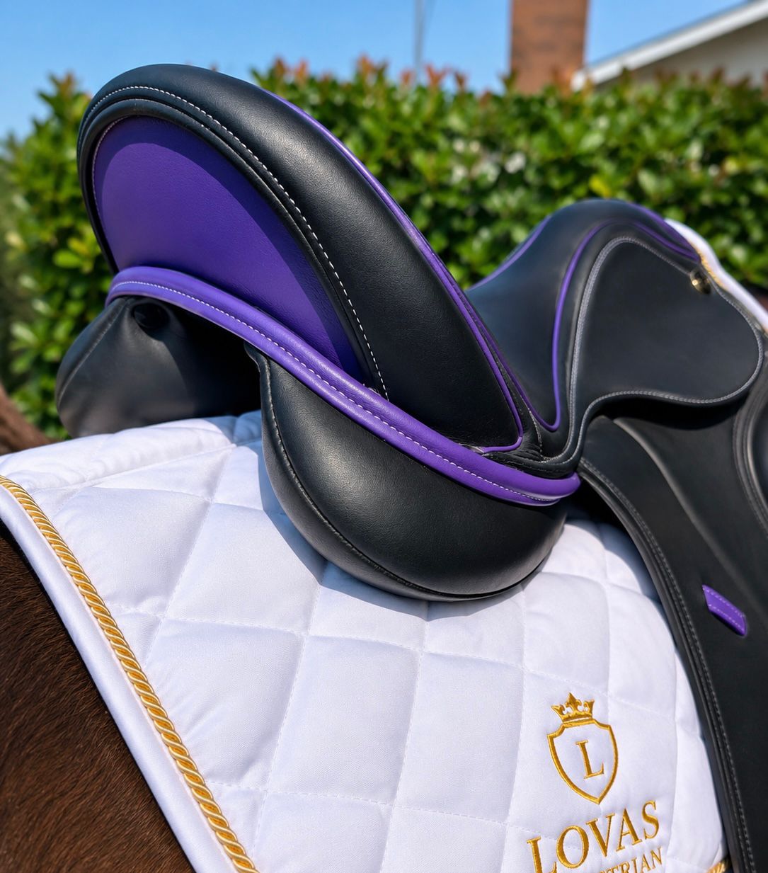 A close-up of a black leather equestrian saddle with purple trim, resting on a white quilted horse pad with gold trim and the LOVAS brand logo, set against a blurred outdoor background of green hedges and blue sky.