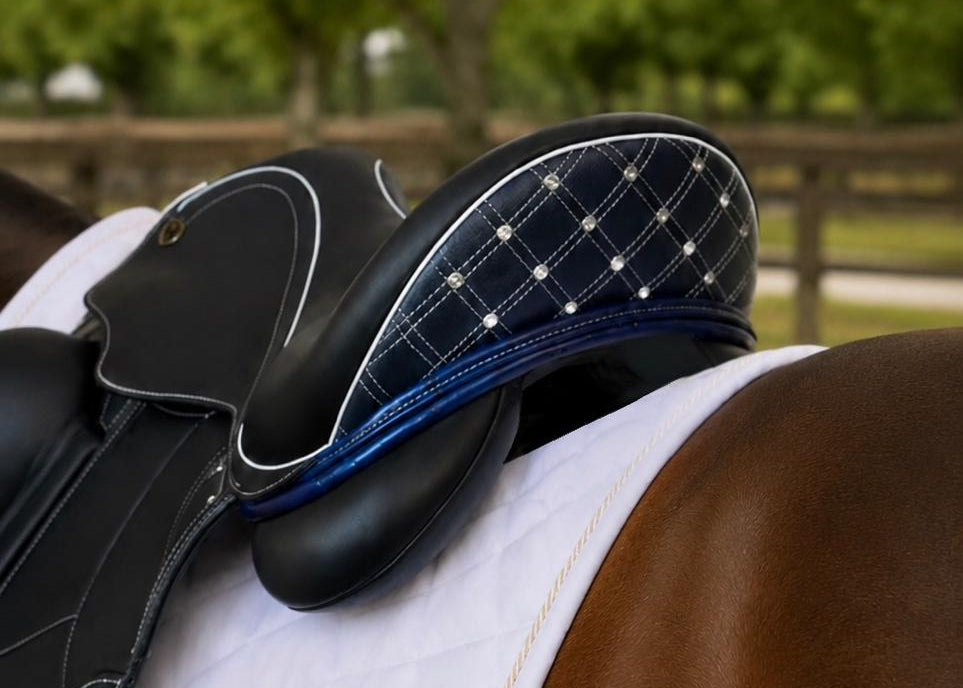 A close-up of an equestrian saddle resting on a horse's back, showing a white quilted saddle pad, a black leather saddle with blue trim, and a navy plaid underwrap with silver rhinestones. The background is a blurred outdoor stable scene with green trees.