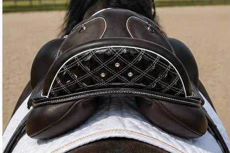 A close-up rear view of a horse wearing a dark brown leather saddle with white contrast stitching and silver studs, set against an outdoor equestrian arena background with trees and sand.