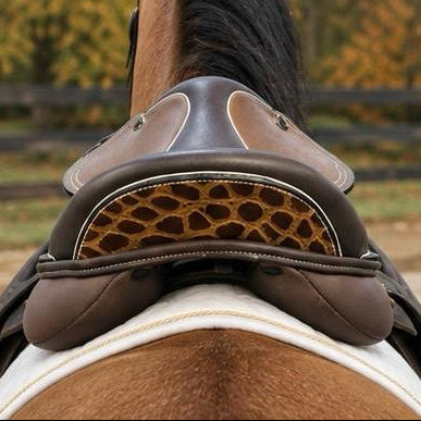A close-up rear view of a horse wearing a brown leather riding saddle with a decorative giraffe-print pattern on the underside, set against a blurred outdoor background of trees and a fence.