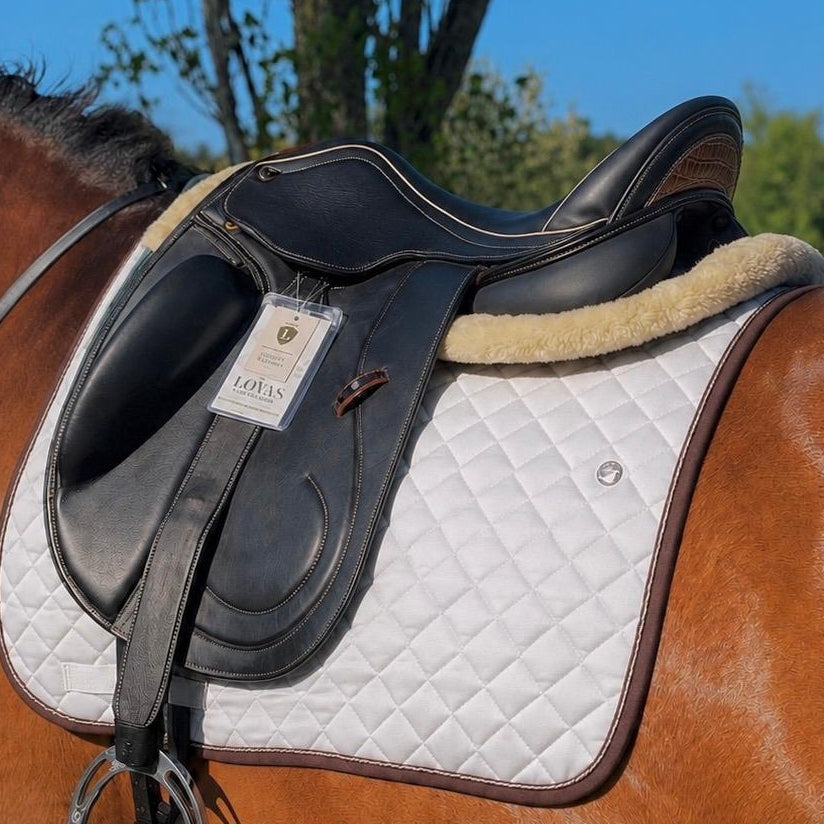 A close-up of a horse saddle with a white quilted saddle pad and tan fleece lining, mounted on a brown horse outdoors under a blue sky.