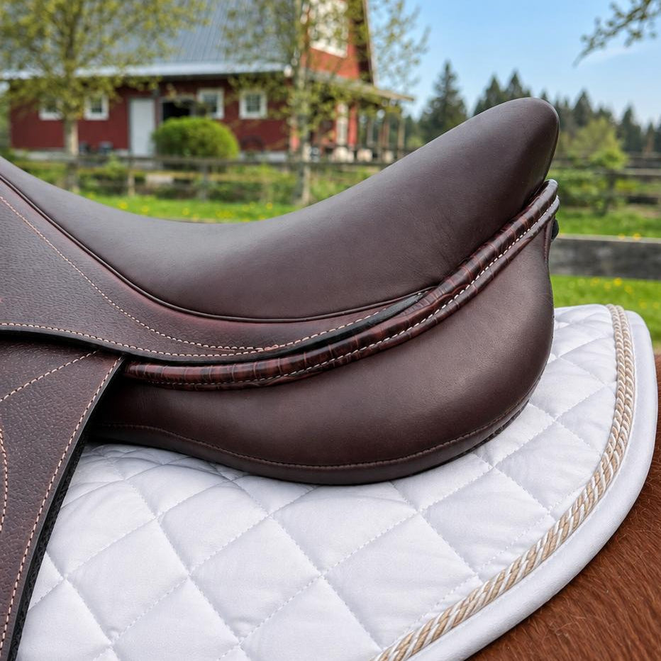 A close-up of a dark brown leather equestrian saddle resting on a white quilted saddle pad, set against a blurred outdoor background of trees, a red barn, and a blue sky.