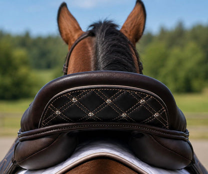 Close-up rear view of a horse wearing a dark brown leather saddle with a quilted diamond-patterned black center panel and silver rhinestone accents, set against a blurred outdoor background of green trees and blue sky.