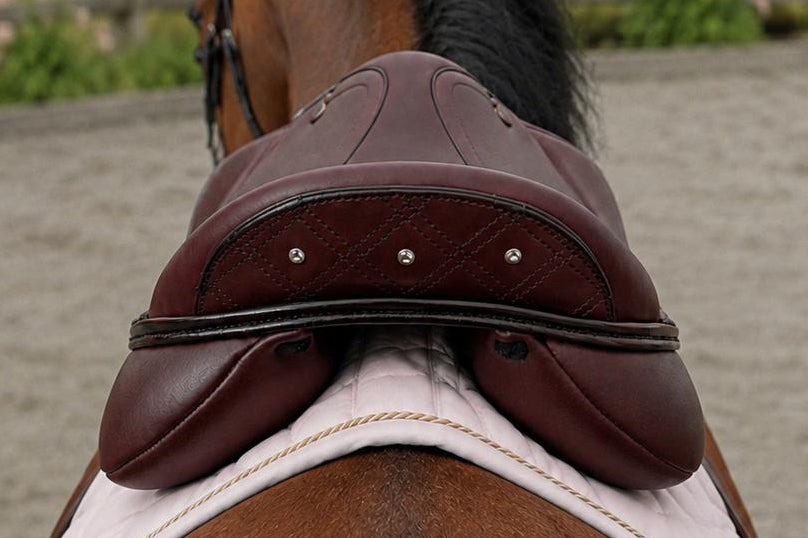 A close-up rear view of a brown horse wearing a dark brown leather saddle and white saddle pad, standing in an outdoor horse arena with a wooden fence and green pasture in the background.
