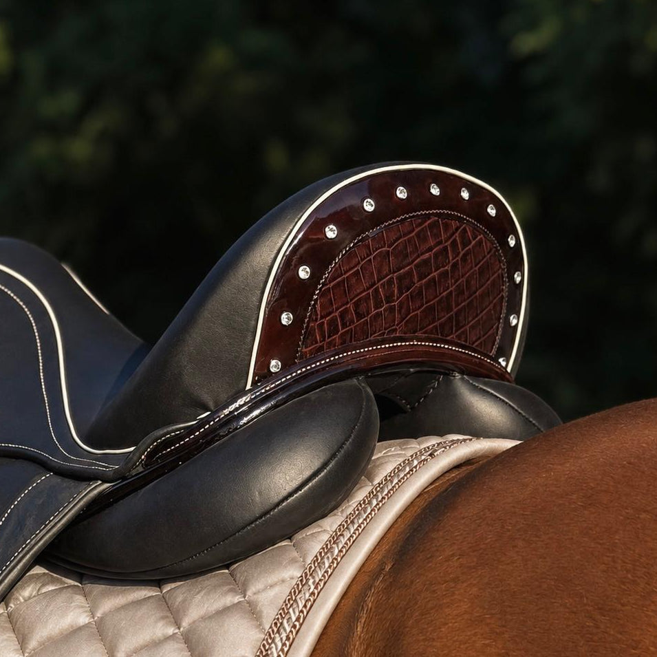 Close-up of a horse saddle resting on a horse's back, showing black leather, dark brown textured leather, silver studs, and a beige quilted saddle pad against a blurred green outdoor background.