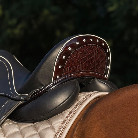 Close-up of a horse saddle resting on a horse's back, showing black leather, dark brown textured leather, silver studs, and a beige quilted saddle pad against a blurred green outdoor background.