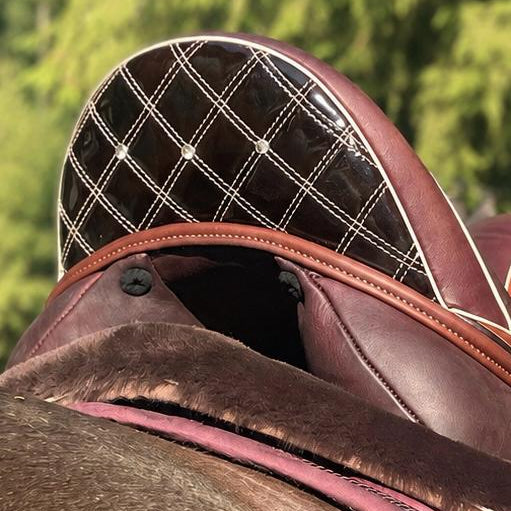 A close-up of a horse saddle with a dark brown leather seat, burgundy velvet saddle pad, and a black diamond-quilted saddle pad insert, shown outdoors on a horse with a blurred forest background.