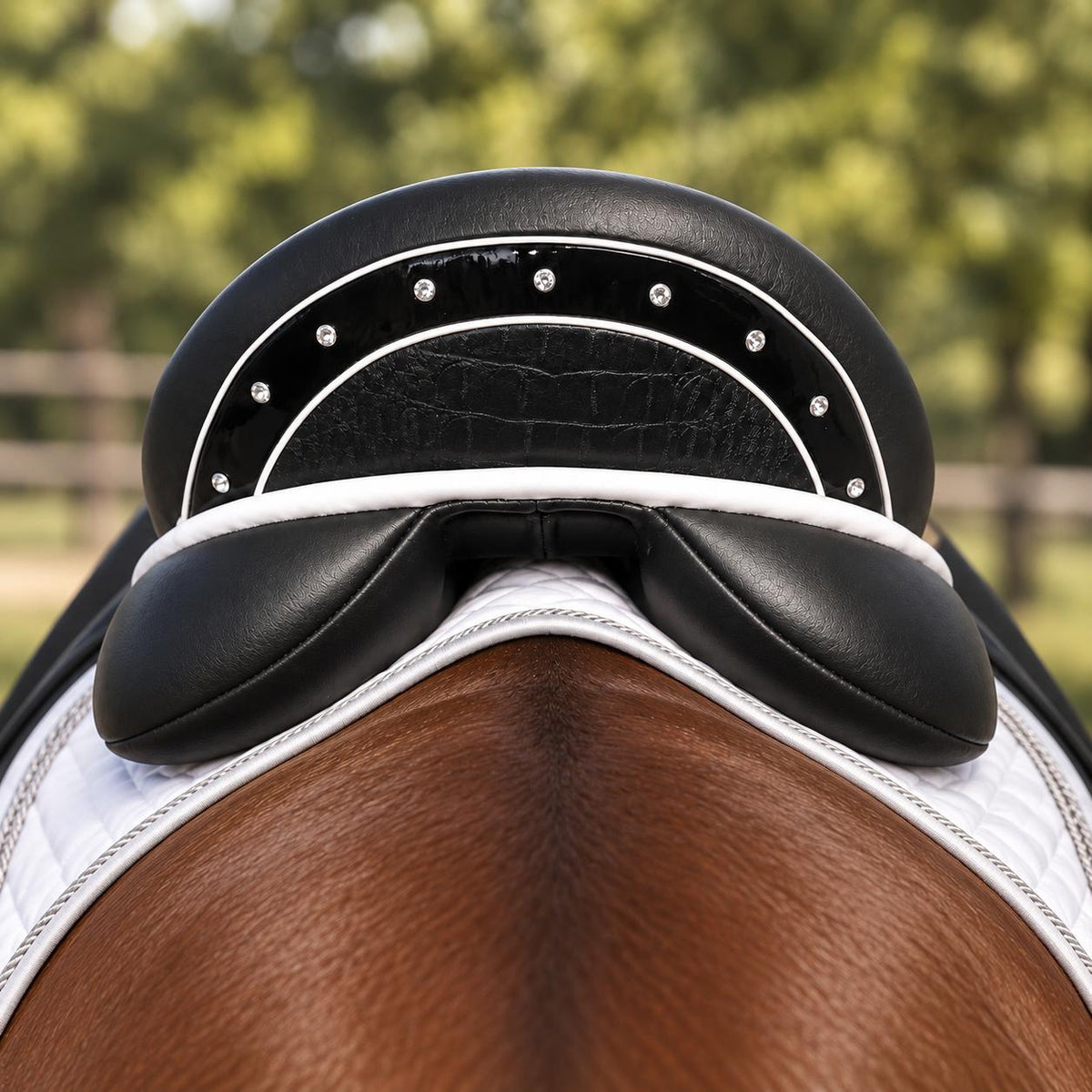 Close-up of a black leather equestrian saddle with white piping and silver studs, positioned on a brown horse's back over a white saddle pad, with a blurred green outdoor background.