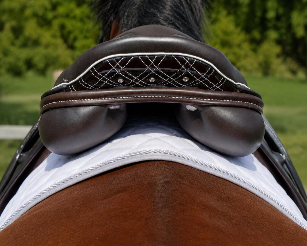 A close-up rear view of a brown horse outdoors, showing a dark brown leather saddle with white embroidered detailing and a white saddle pad against a blurred green forest background.